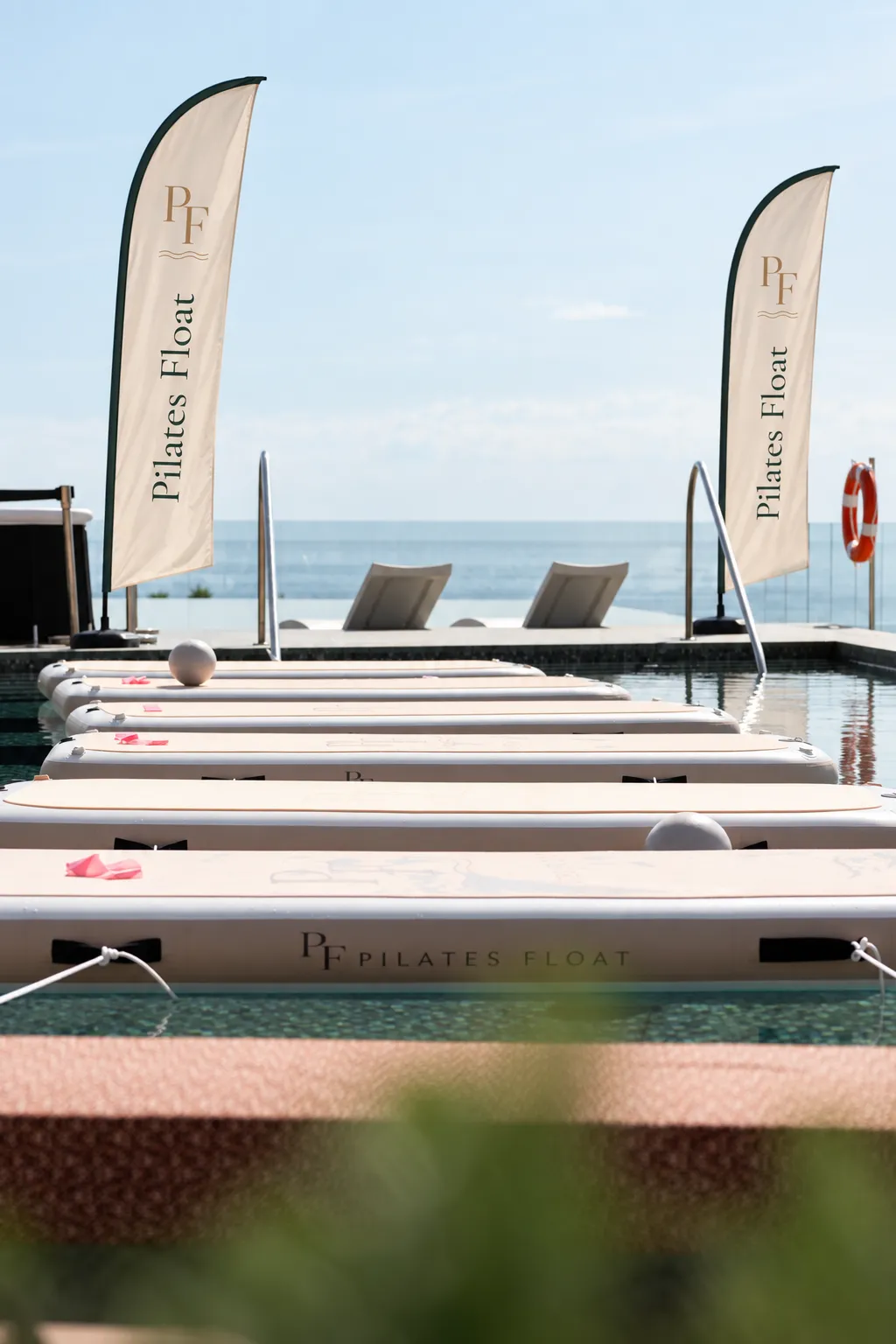 Branded Pilates Float boards lined up on a hotel rooftop pool, flags against the sea